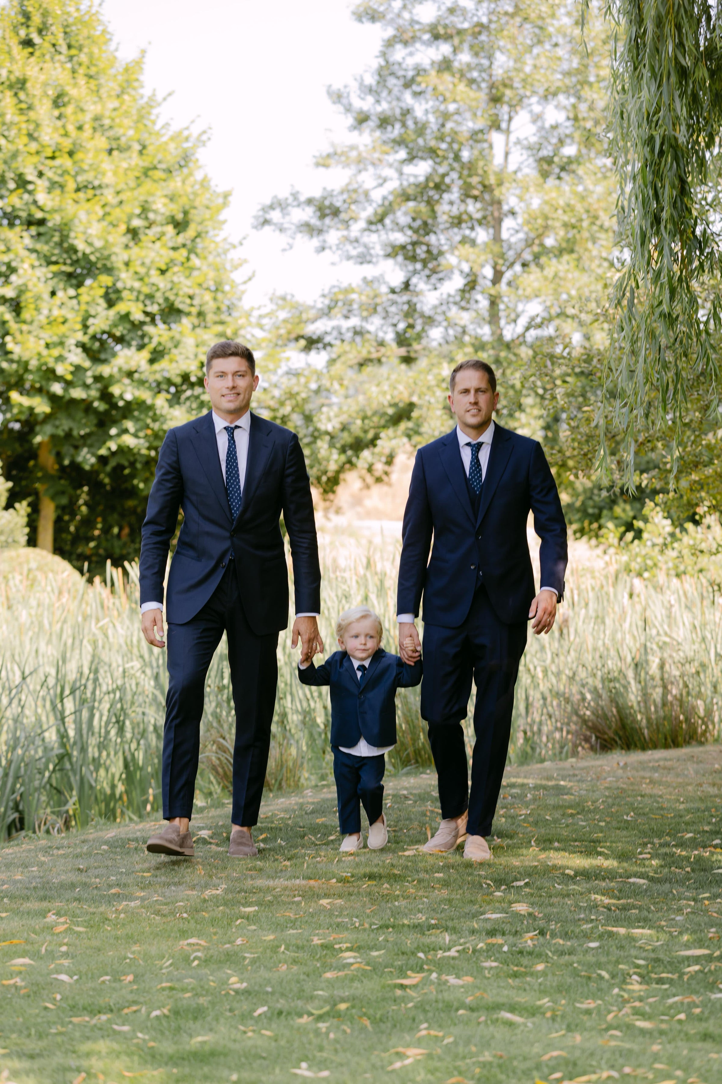 Two men in suits walking with a child on a grassy path with trees in the background