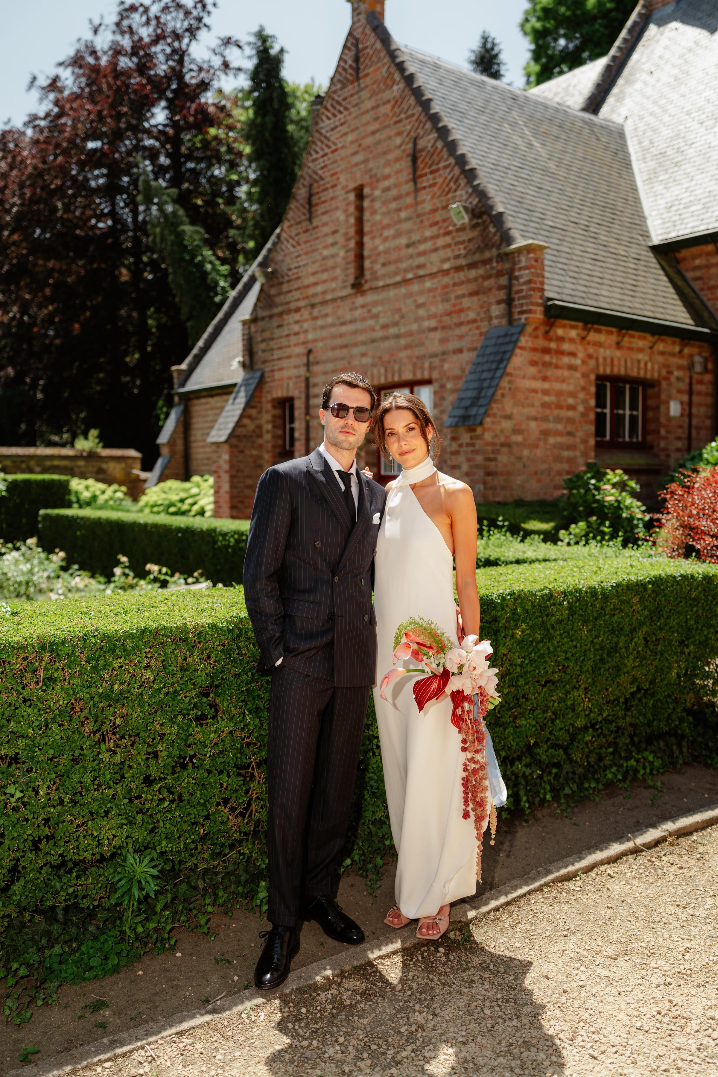 Man and woman in formal attire standing in front of a brick building with greenery.