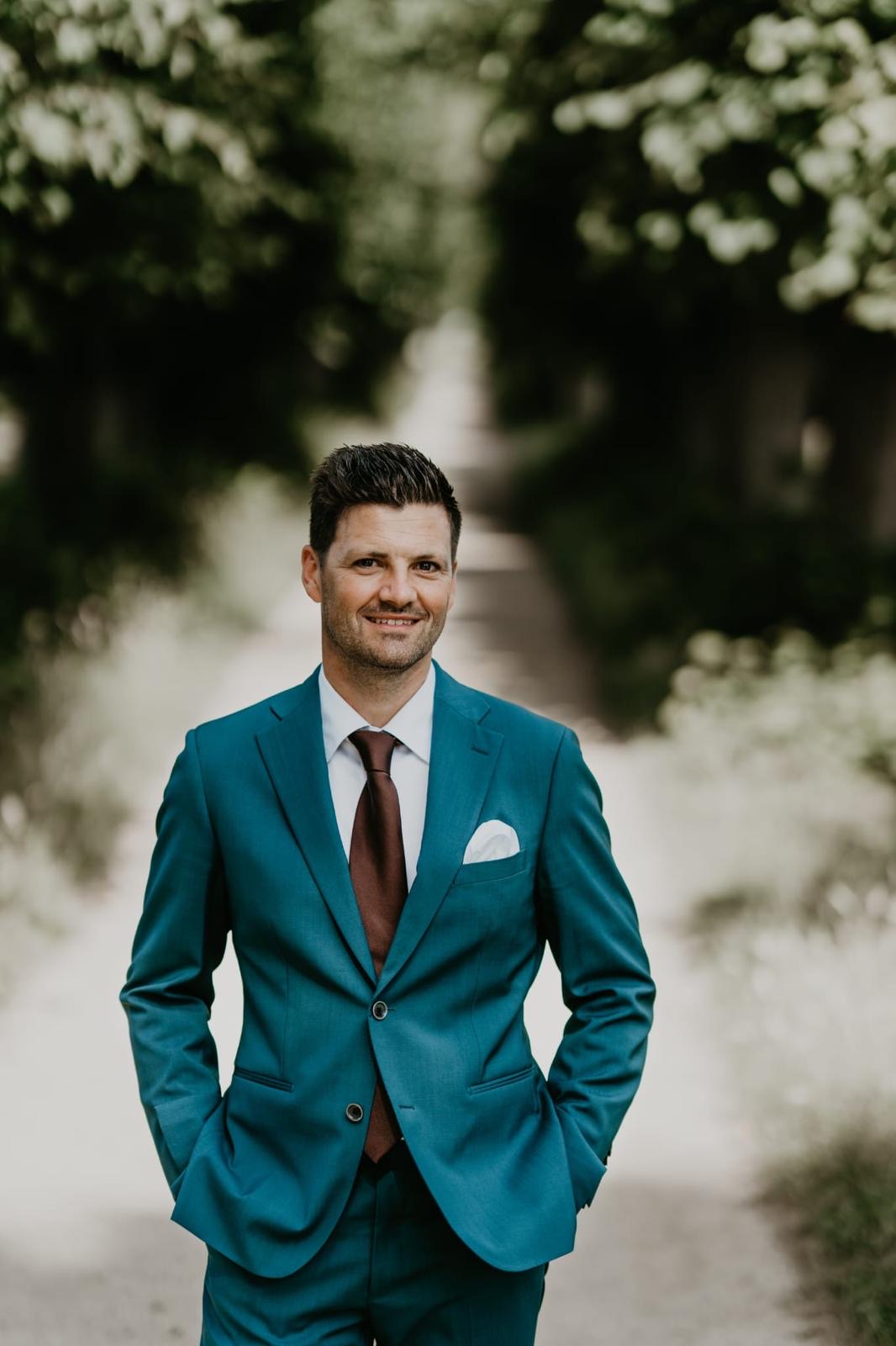 Man in a teal suit with a red tie standing outdoors with trees in the background