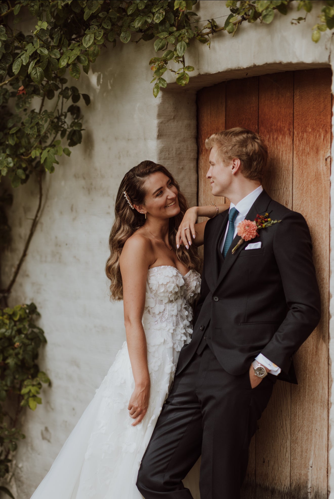 Couple in wedding attire standing against a rustic wall with greenery.