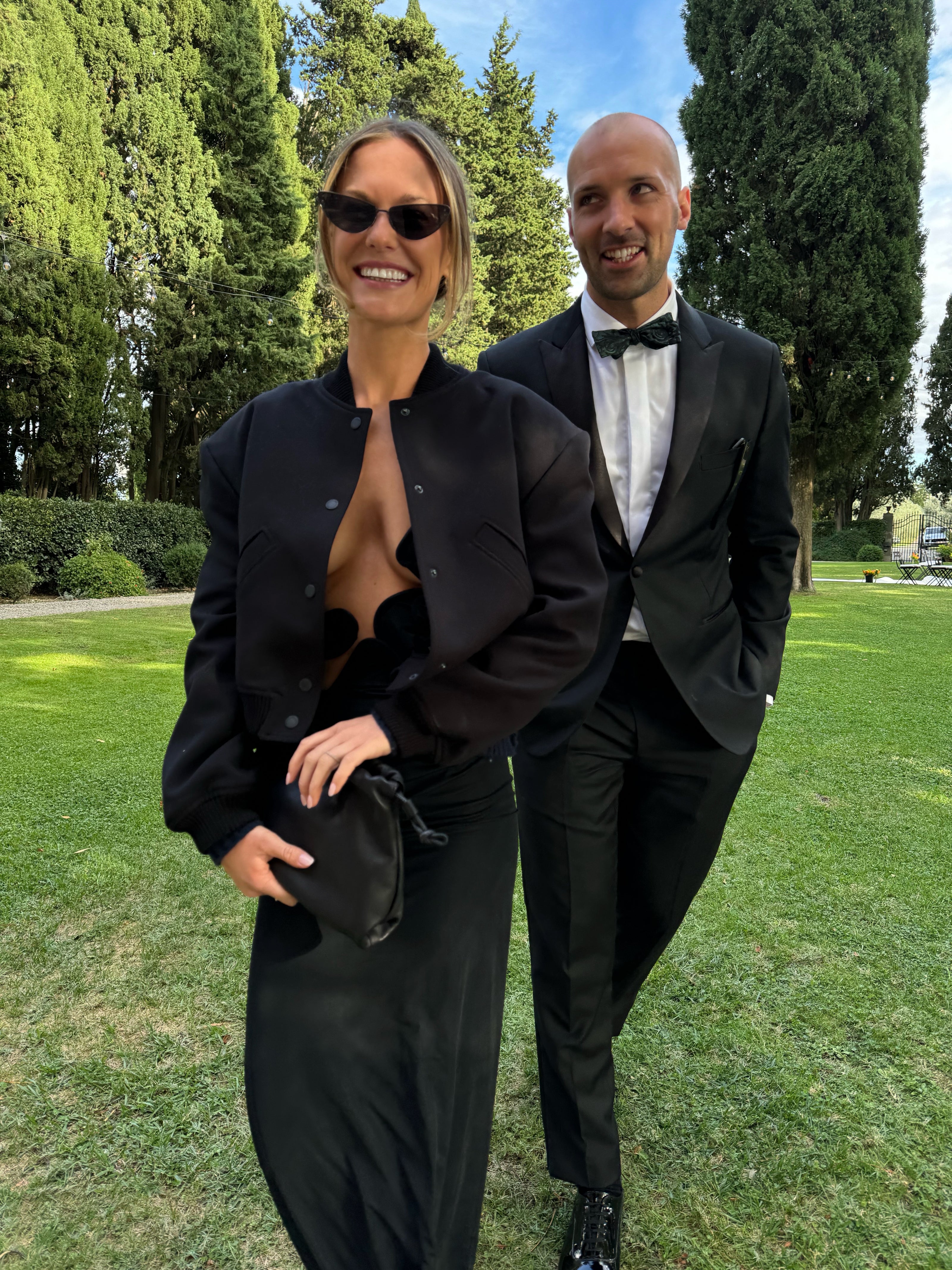 Two people in formal attire standing on a grassy area with trees in the background
