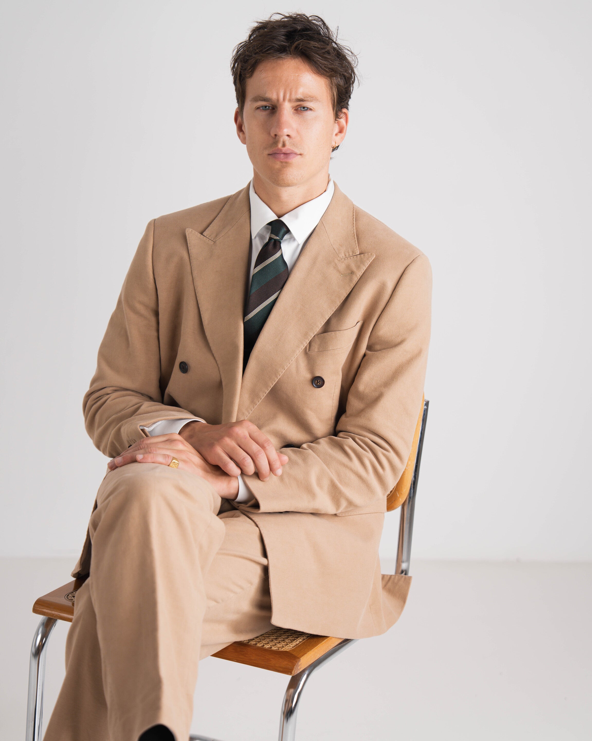 Man wearing a beige trench coat sitting on a chair against a plain background