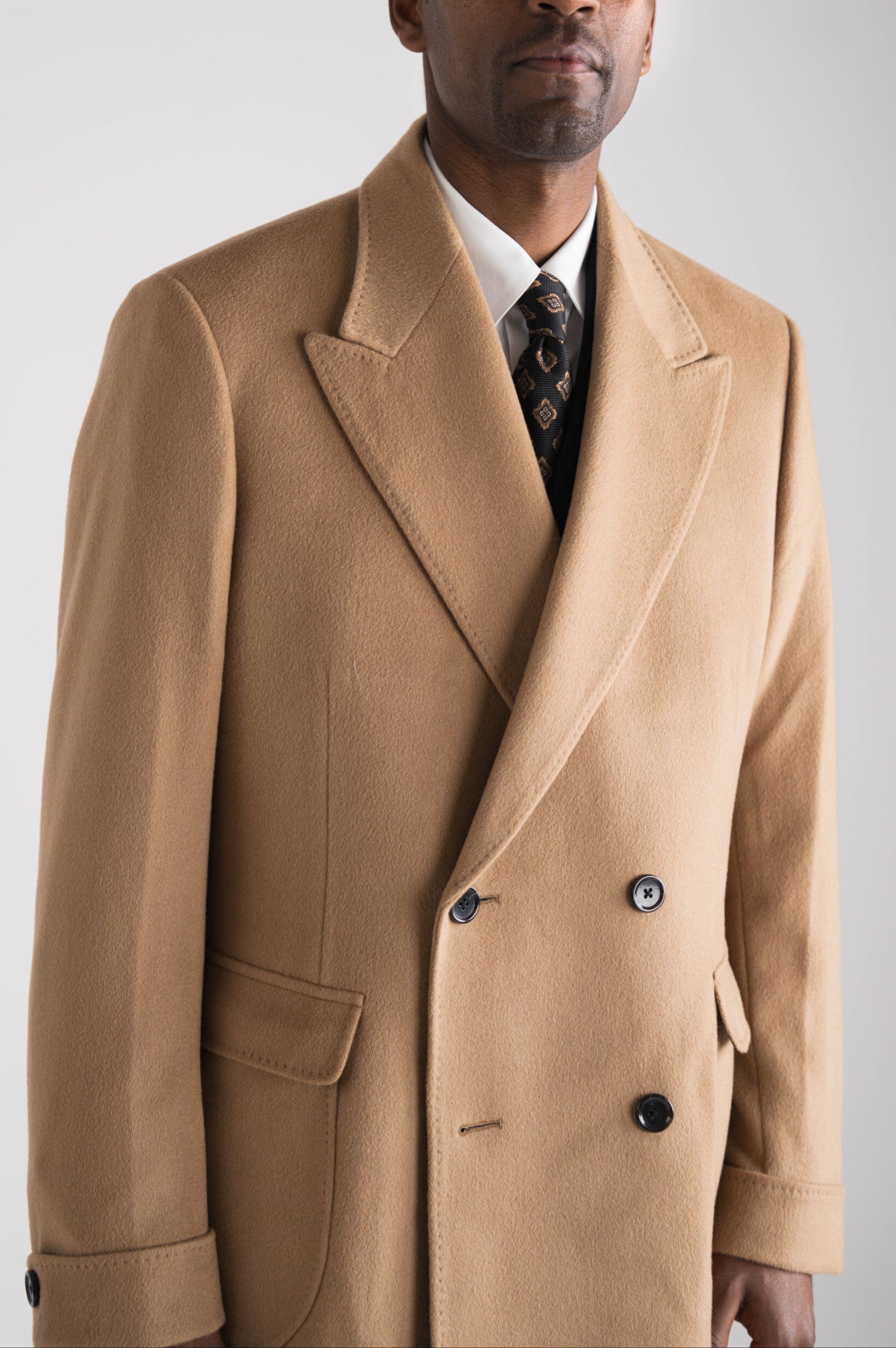 Man in beige overjas op maat tegen een neutrale achtergrond