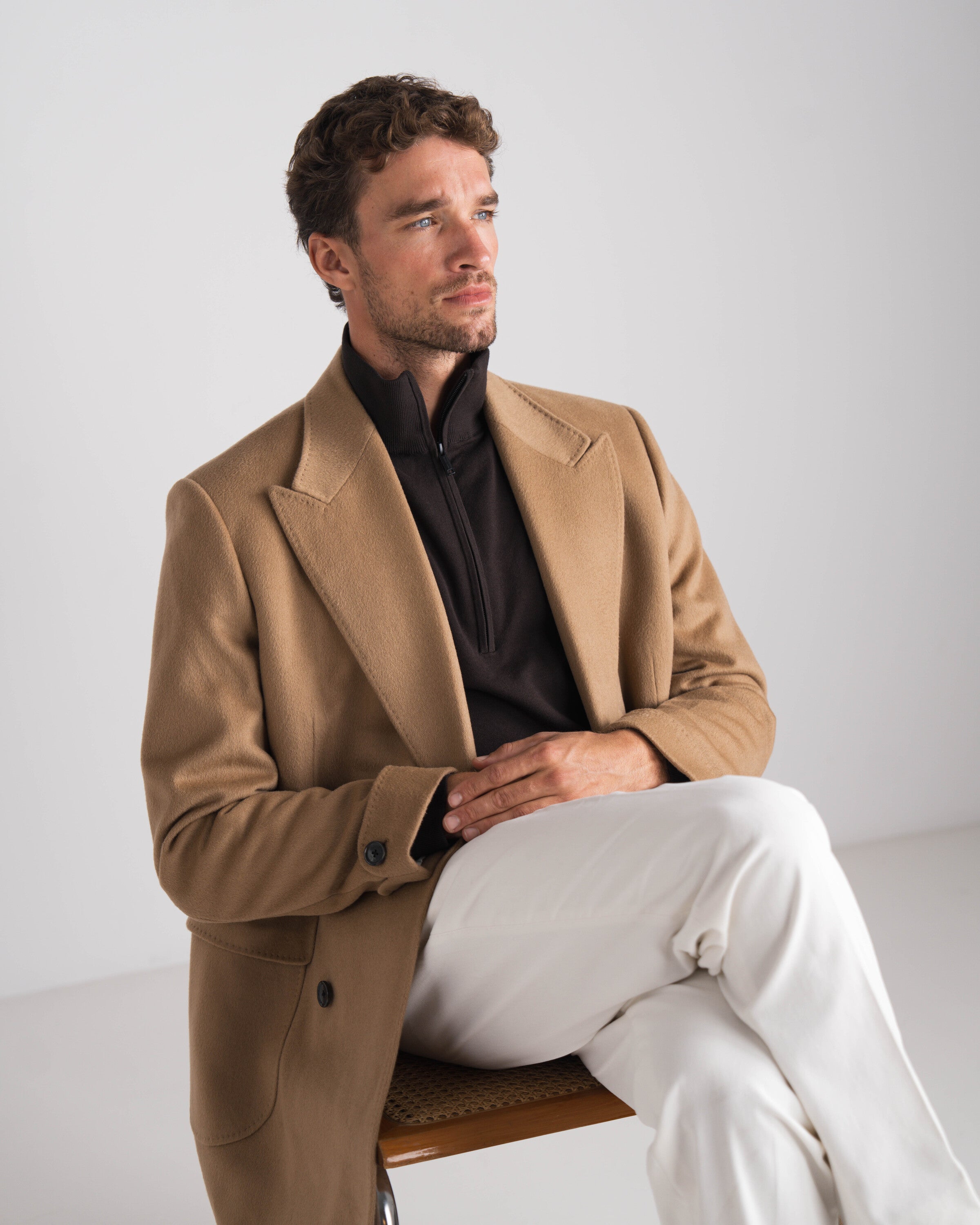 zittende man met bruine trui en camel overcoat en witte broek