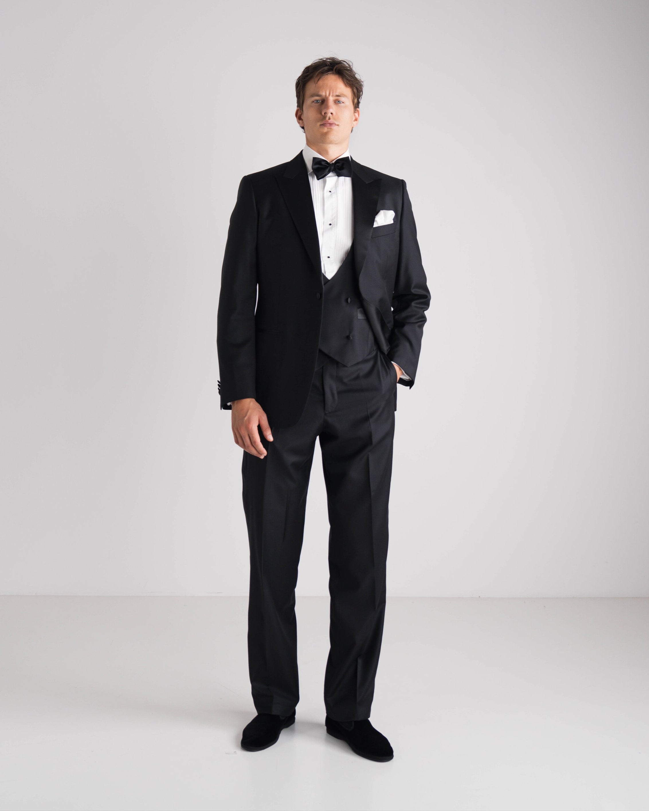 Man wearing a black tuxedo with a white shirt and bow tie on a plain background