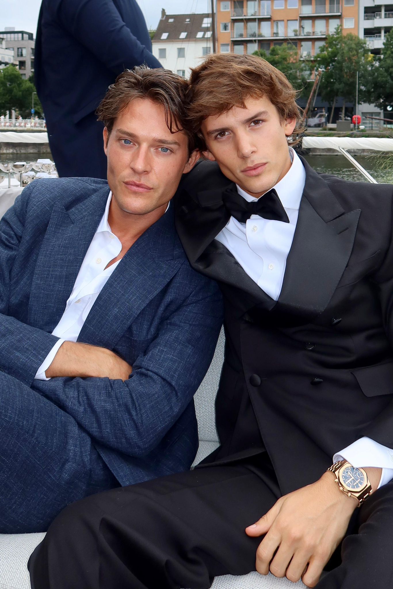 Two men in formal attire sitting together with a cityscape background. Cesar Casier and Conrad Janssens