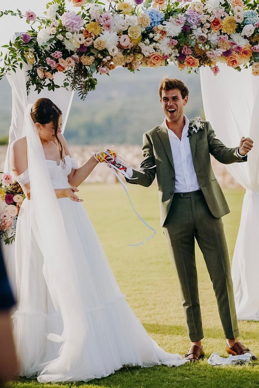 Couple in wedding attire standing under a floral archway on a grassy field.