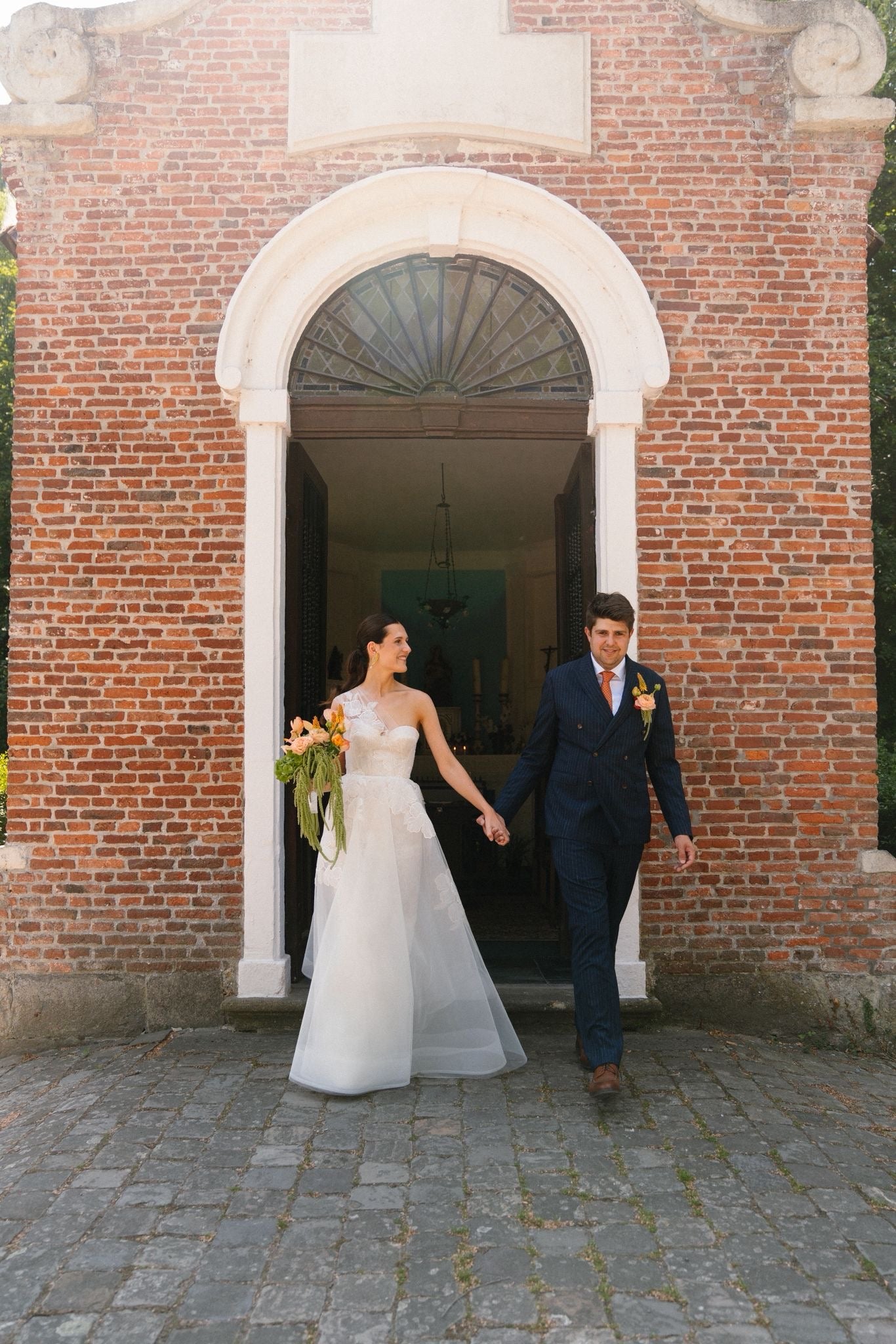 Couple in wedding attire standing in front of a brick building with an archway.