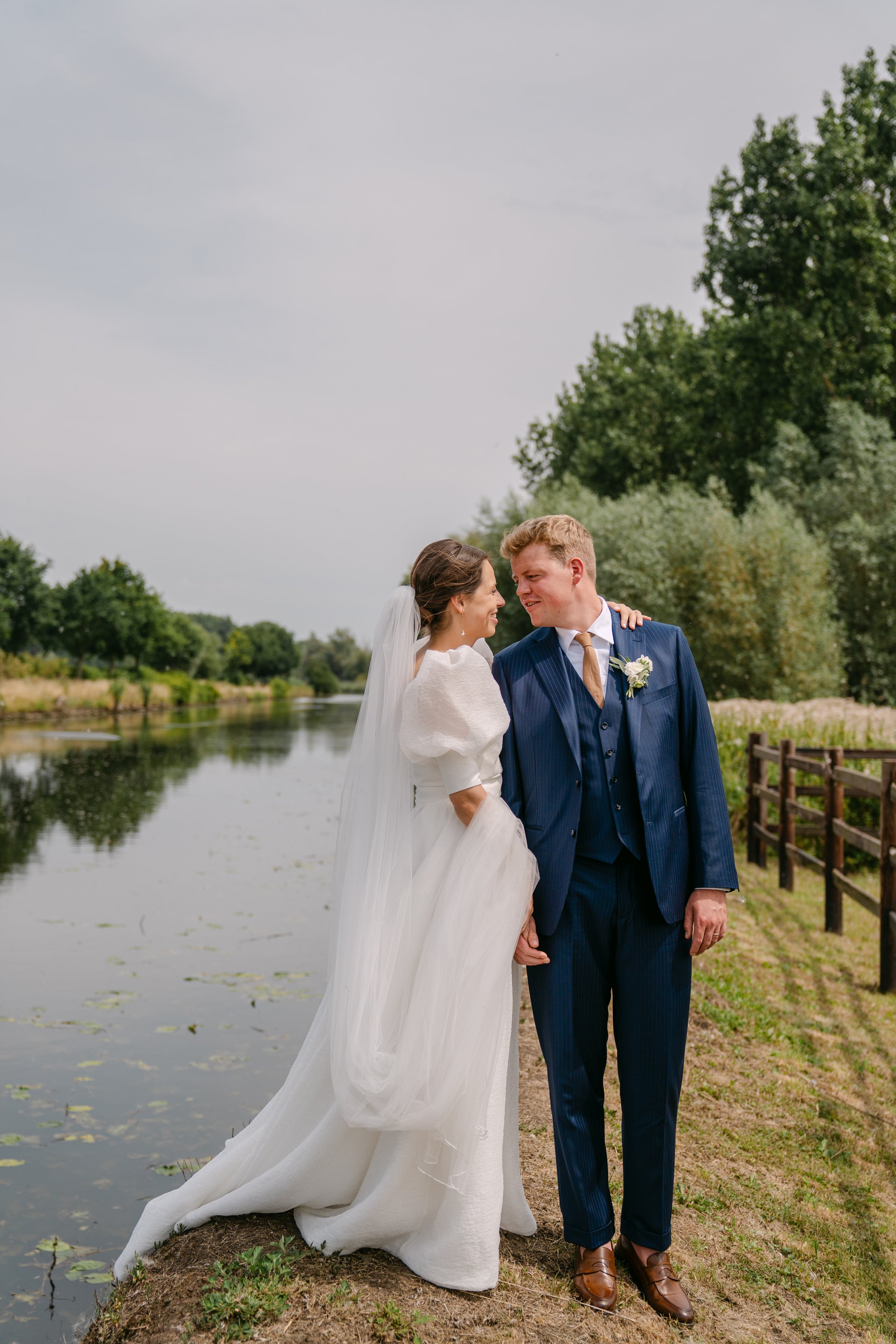Couple in wedding attire standing by a pond with trees in the background