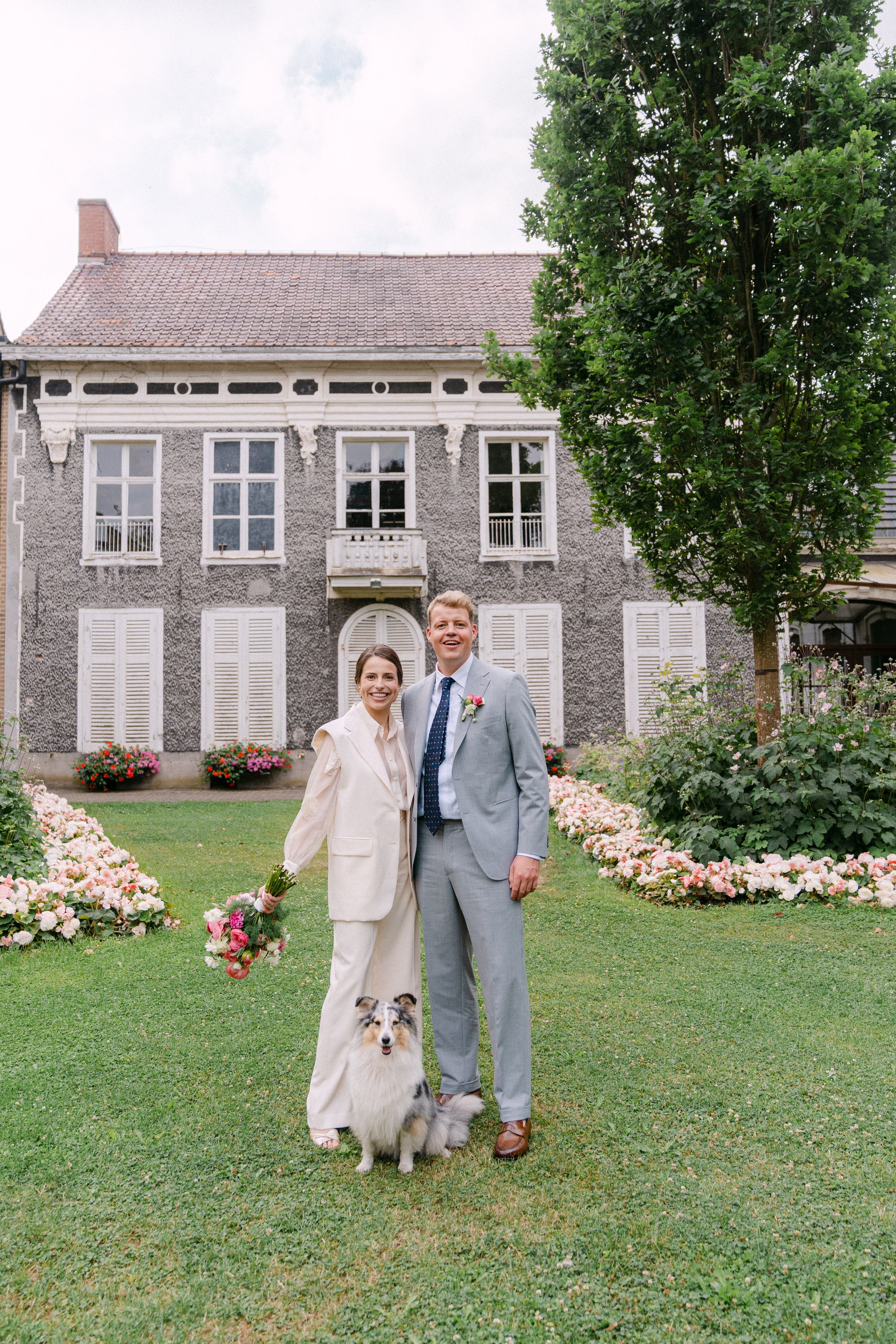 Couple standing on a grassy lawn with a dog, in front of a building with decorative flowers.